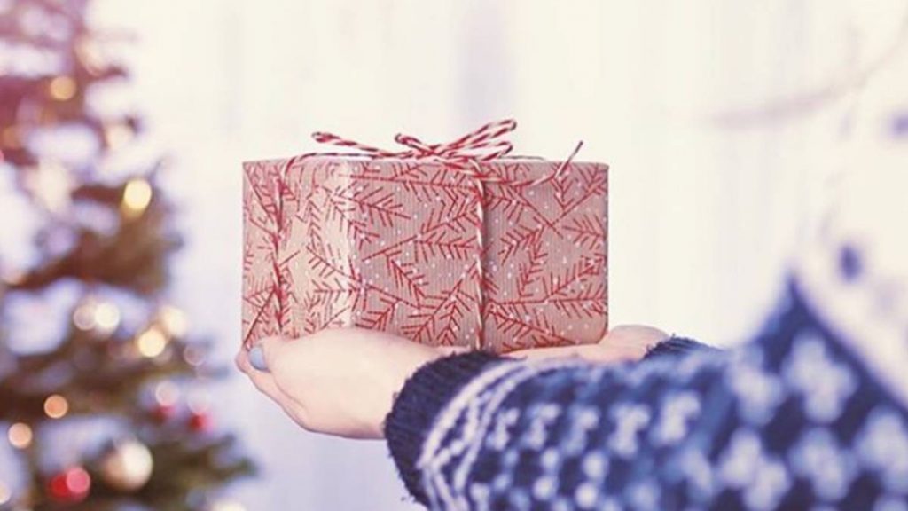 La mano de una mujer sosteniendo una caja de regalo frente a un árbol de Navidad.